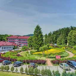 Tanie sanatorium prywatnie: budynek z czerwonym dachem, parking z samochodami i piękny ogród z kolorowymi kwiatami.