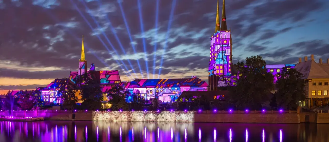 Ostrów Tumski we Wrocławiu nocą, iluminowany kolorowymi projekcjami. Trasa spacerowa wzdłuż rzeki odbija światła.